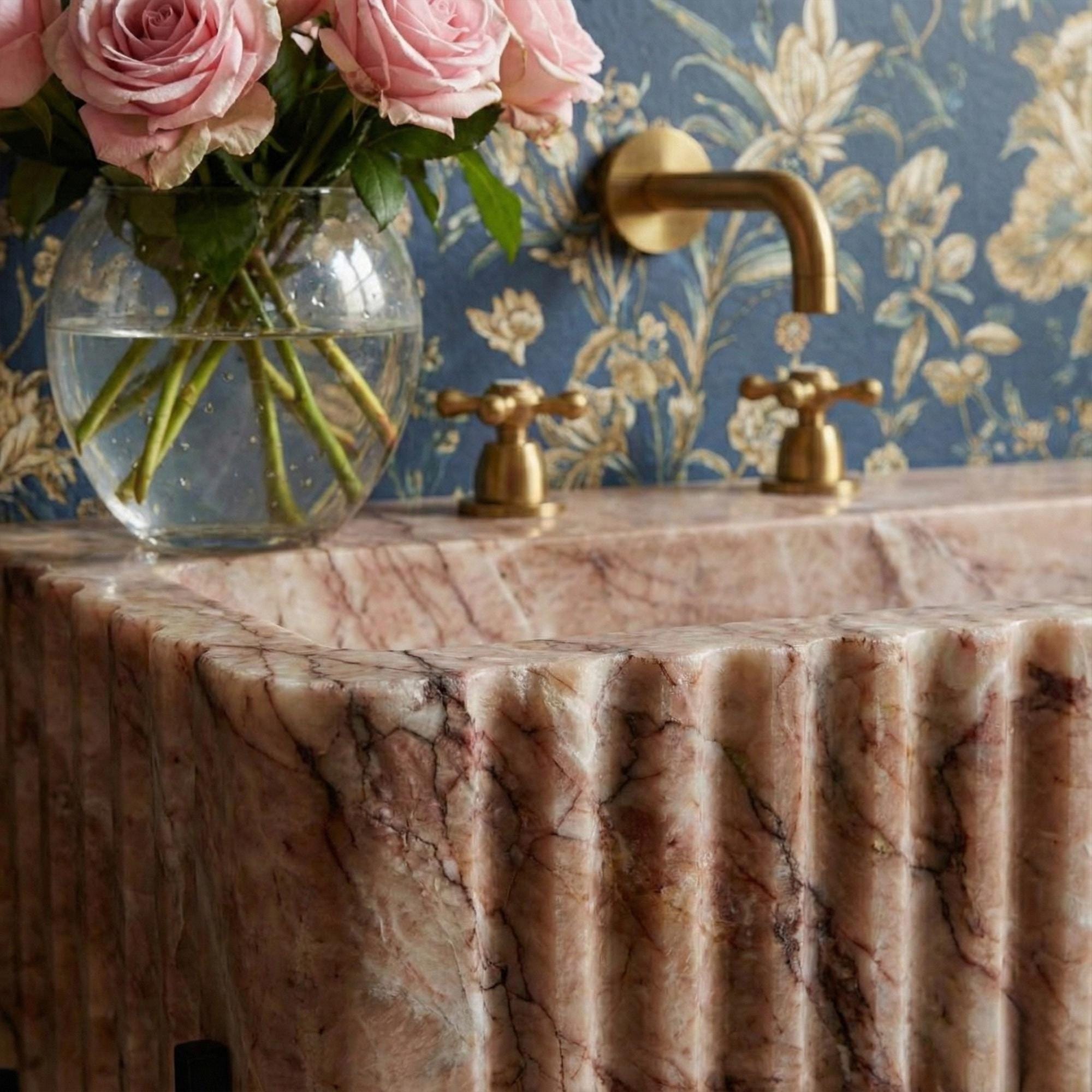 Statement fluted console sink crafted from pink marble, featuring black metal frame and floral wallpaper backdrop.