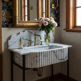 St. Moritz white marble fluted sink with scalloped integrated backsplash and luxurious brass fixtures, mounted on a black console stand.